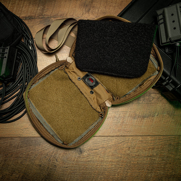 An open tactical pouch with velcro padding is placed on a wooden surface. Nearby are coiled cables and parts of another black bag or equipment, partially visible in the frame. The pouch appears to be intended for carrying gear or accessories such as a Premier Body Armor SOE FUPA Level IIIA Insert.