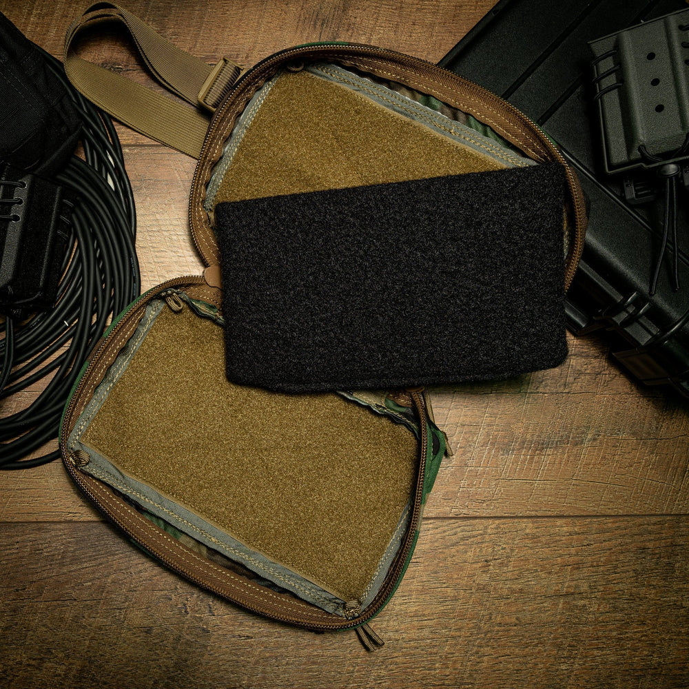 A small, brown SOE FUPA XL Fanny Pack is open on a wooden surface, revealing yellowish-brown interior compartments crafted from durable ballistic material. A Premier Body Armor SOE FUPA XL Level IIIA Insert rests atop the pack. Surrounding it are various cables and a black plastic case.