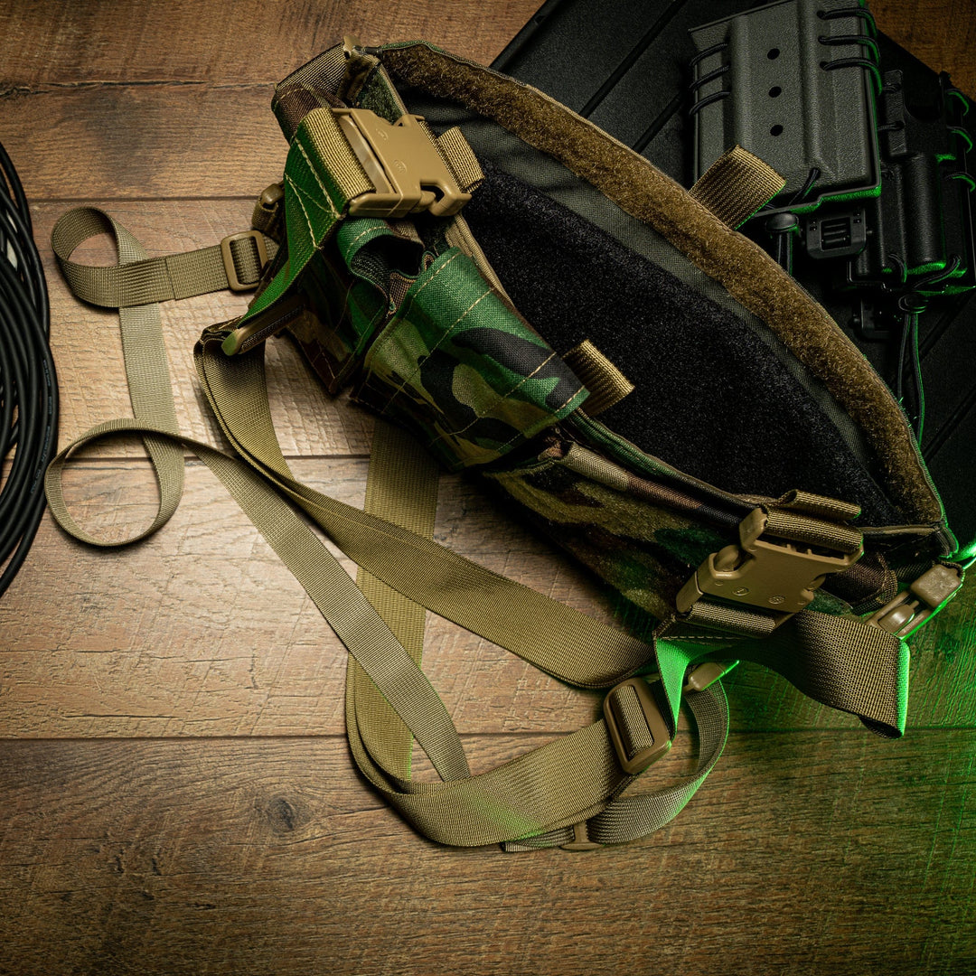 Military gear, including a camouflage-patterned utility belt with tan straps and buckles, is laid out on a wooden floor. Adjacent to it are coiled black cables, a custom fitted black case, and a Premier Body Armor SOE Micro Rig Level IIIA Insert, all illuminated by a green overhead light.