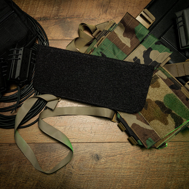 A Premier Body Armor SOE Micro Rig Level IIIA Insert, featuring a camouflage pattern and multiple pouches, is laid out on a wooden surface with a black rectangular patch placed on top. To the left, coiled black cords are visible next to beige straps extending from the custom-fitted micro rig.