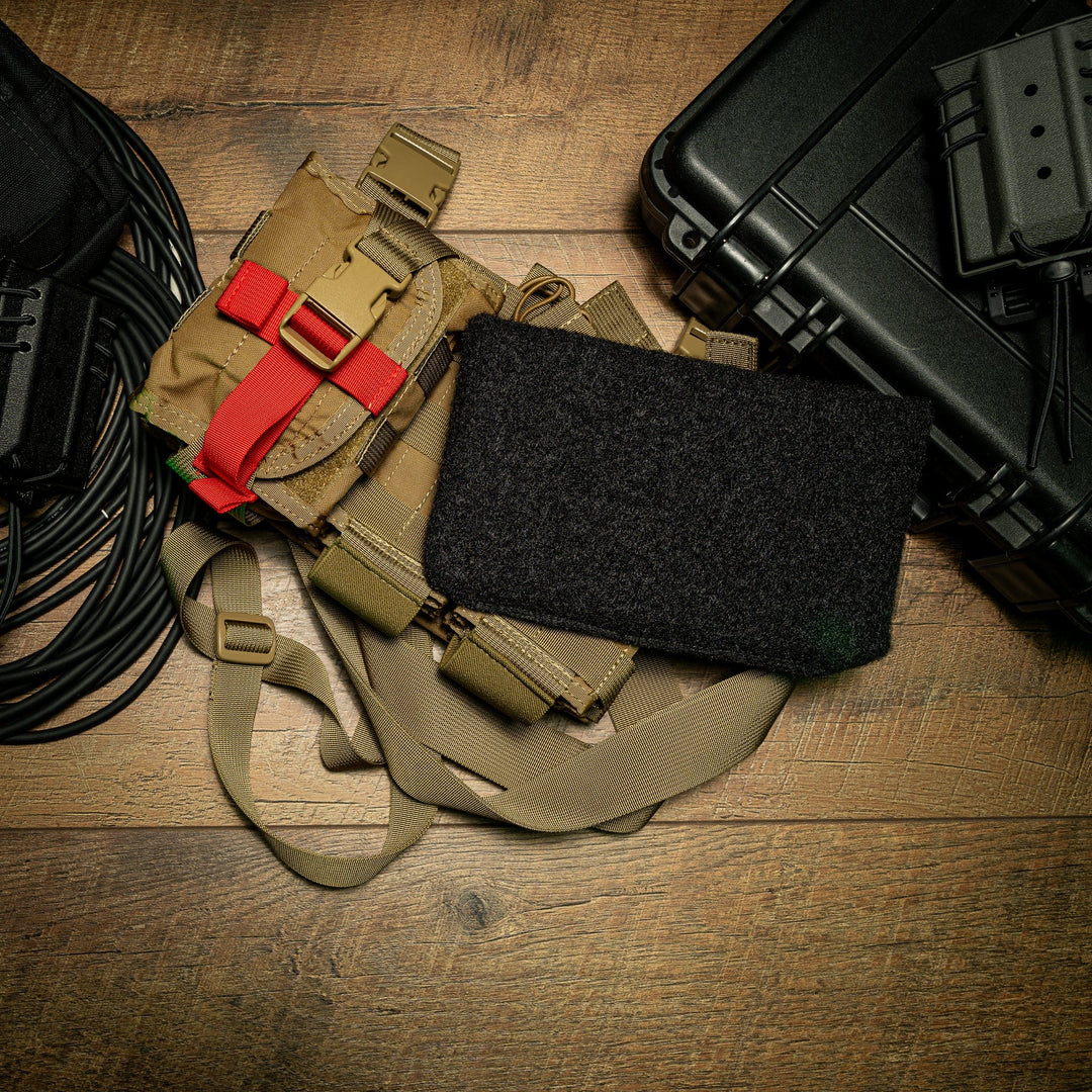 A flat-lay image of tactical gear on a wooden surface. Items include a tan harness with red straps, a black utility case, a coiled cable, and an SOE Stacked Micro Rig next to Premier Body Armor's Level IIIA Insert crafted from ballistic material.