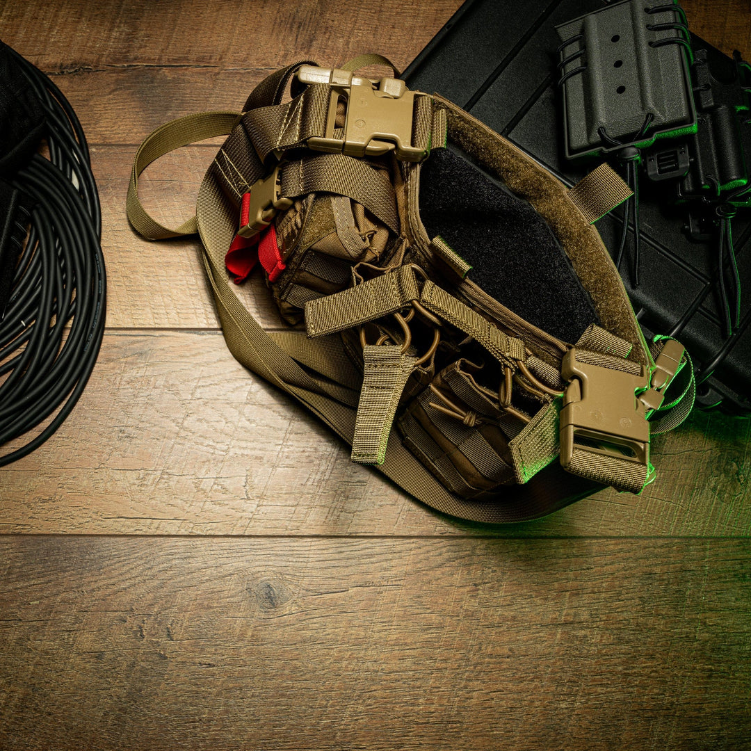 A tactical gear setup on a wooden surface includes a tan harness with multiple straps and buckles, a black coiled rope to the left, a black case on the right, and a Premier Body Armor SOE Stacked Micro Rig Level IIIA Insert partially visible on the far right.