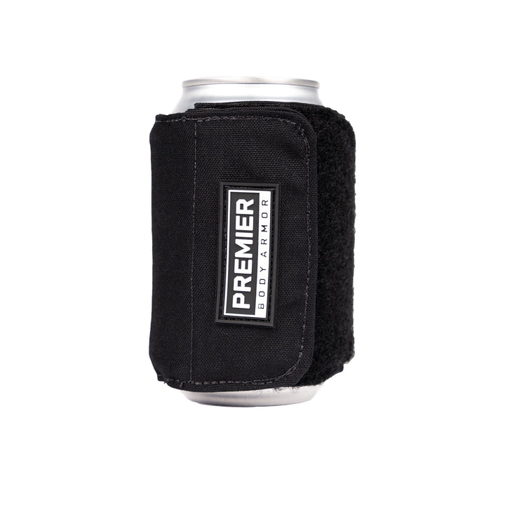 A can of beverage is encased in a black, textured fabric sleeve with a Velcro closure. The sleeve, labeled "Premier Body Armor" on the front tag, features ballistic fibers for added durability. This is known as the Bulletproof Can Cooler from Premier Body Armor.