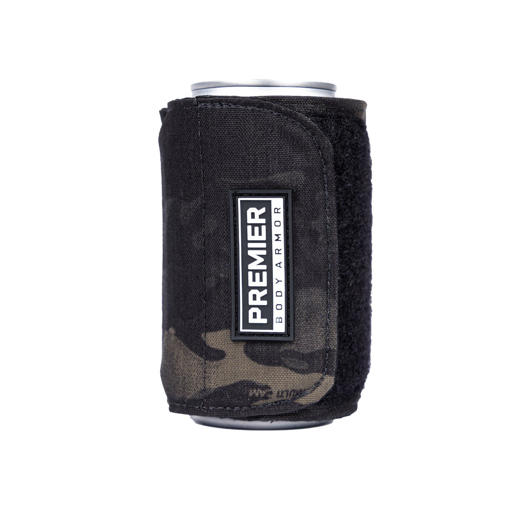 A cylindrical object, likely a beverage can, encased in a black camouflage-patterned sleeve made from ballistic material with a Velcro closure. A white label on the sleeve reads "Premier Body Armor." This innovative product is known as the Bulletproof Can Cooler.