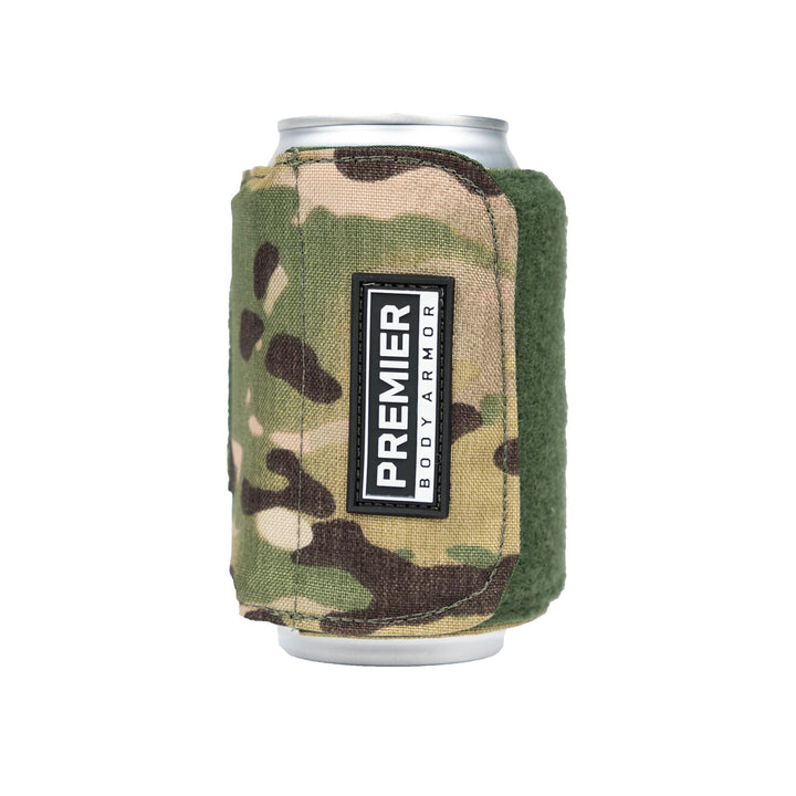 A beverage can is partially covered by the Bulletproof Can Cooler, a camouflage-patterned fabric holder with a green edge from Premier Body Armor. The cooler features a rectangular label with the text "PREMIER BODY ARMOR" and incorporates ballistic fibers for NIJ - Level II protection.