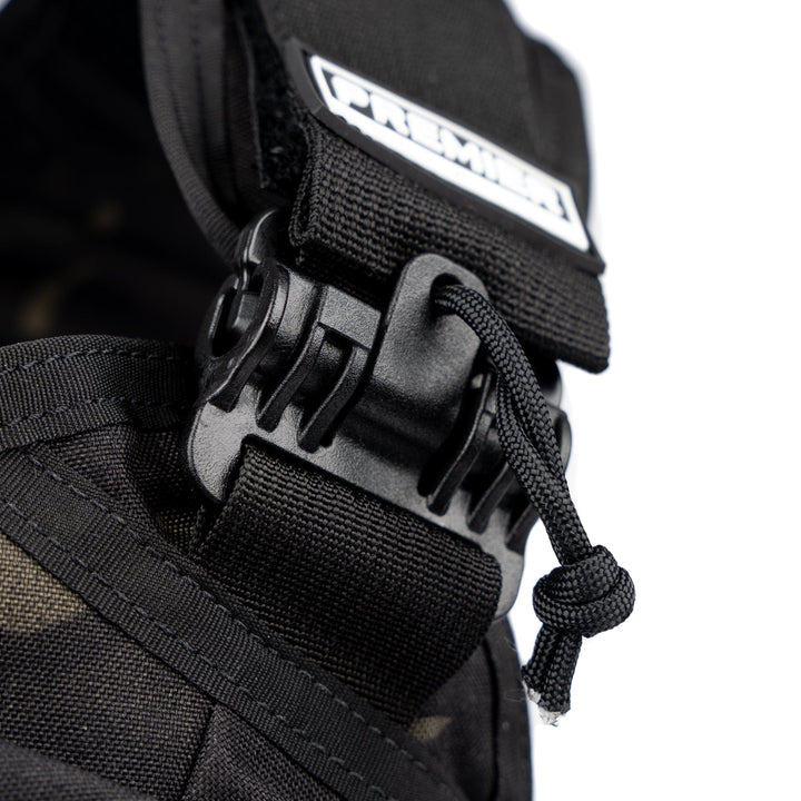 Close-up of a black carabiner-style buckle used to secure straps on the Core Mission Plate Carrier with Level IIIA Cummerbund Armor. The buckle is attached to a fabric strap, with a looped cord hanging next to it. The word "Premier" is visible on a stitched label near the top, highlighting its durability.