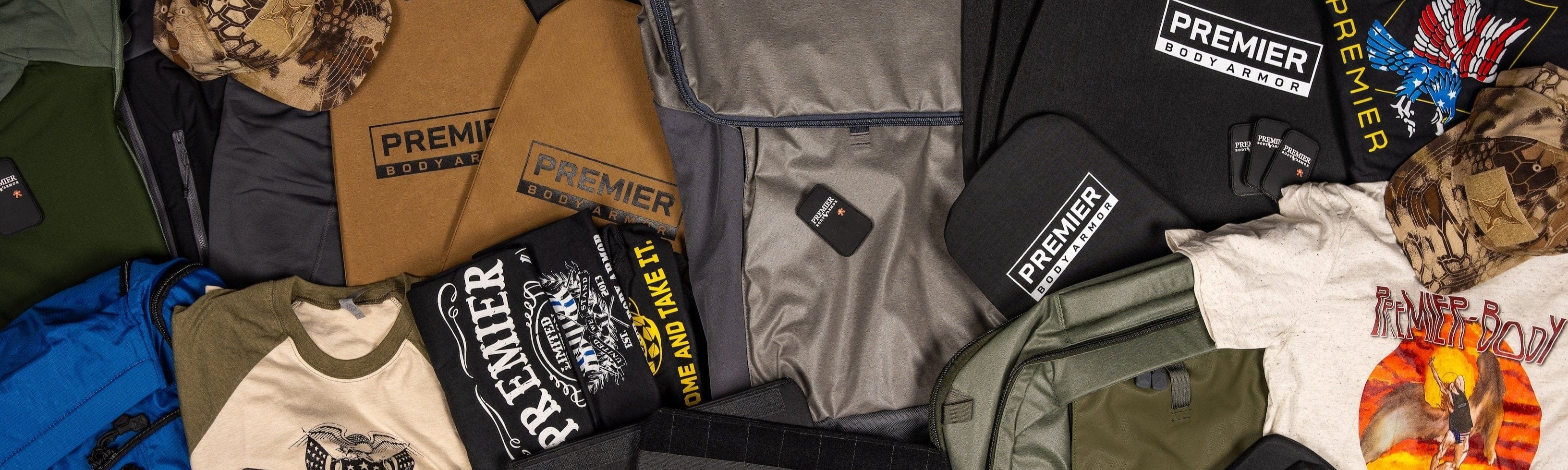 A variety of Premier Body Armor branded bags, shirts, hats, and patches are arranged on a flat surface, showcasing different colors, logos, and designs.