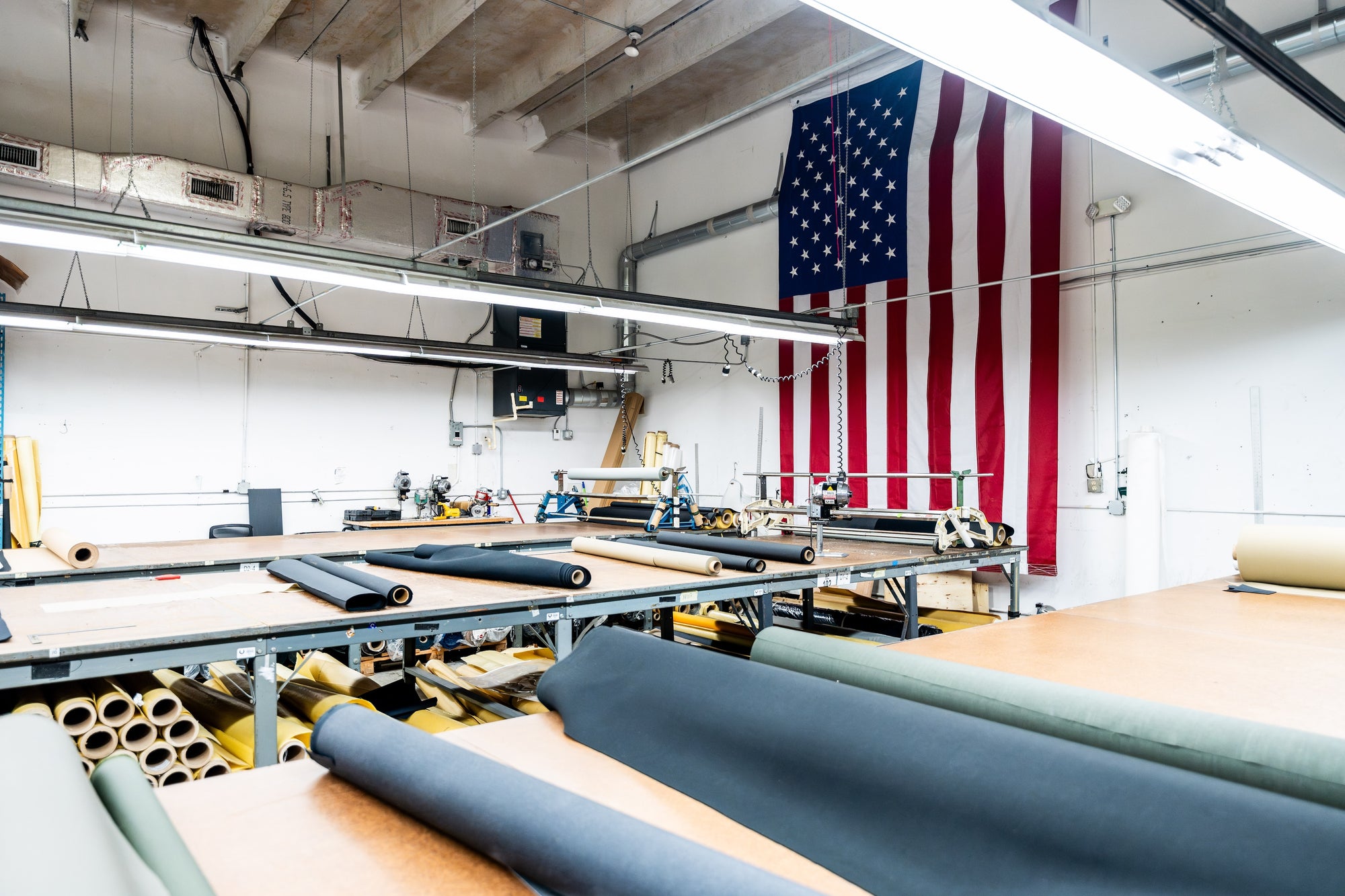 An industrial workspace with large tables covered in various rolls of fabric. Sewing machines are visible, and a large American flag hangs on the wall. Fluorescent lights illuminate the room.