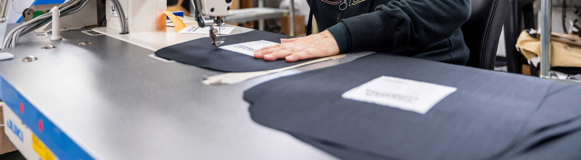 A person operating a sewing machine is stitching fabric pieces labeled with tags. The work is being performed on a metallic table, and the individual is wearing a dark-colored garment.