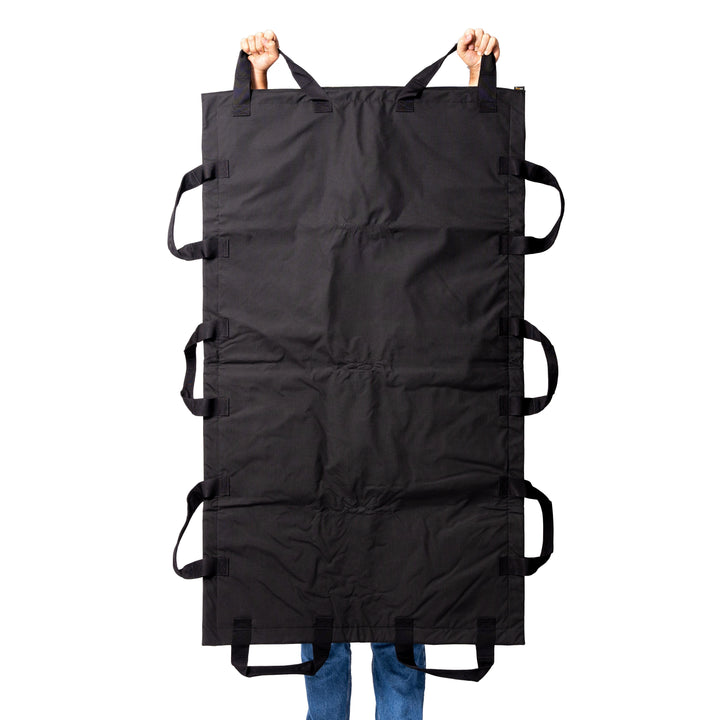 A person is holding up the Fortis Ballistic Blanket by Premier Body Armor, a large rectangular item with multiple side handles. Made from durable material, it looks sturdy and protective. The person's feet are visible, clad in blue jeans and brown shoes.