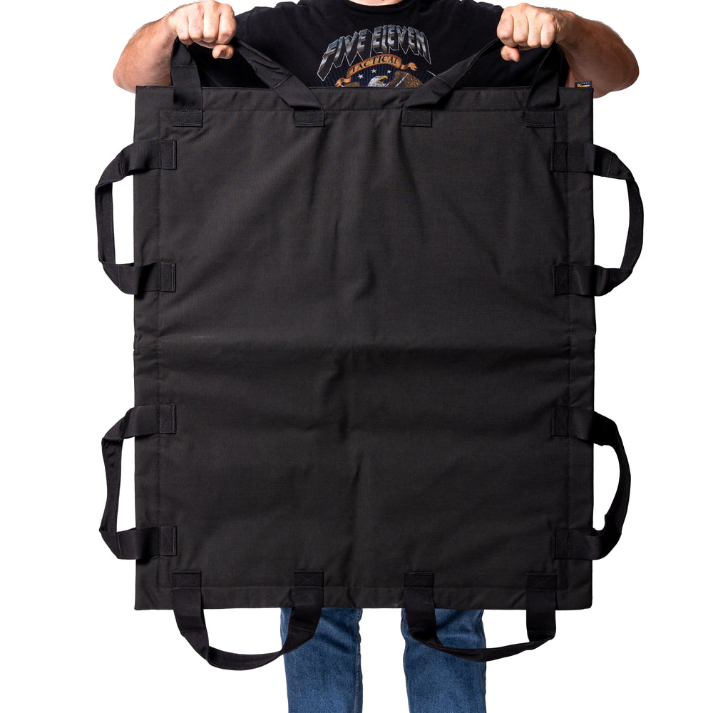 A person is holding a large, rectangular black fabric item with multiple handles along its edges, resembling the Fortis Ballistic Blanket by Premier Body Armor. The person's face is not visible, and they are wearing blue jeans and a black graphic t-shirt against a plain white background.