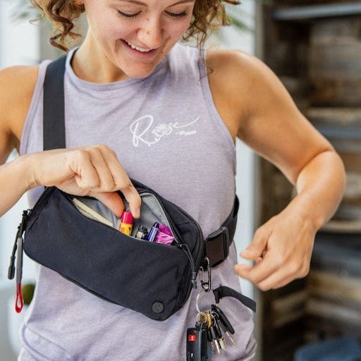 A person with curly hair wearing a sleeveless top opens the Vertx Everyday Fanny Pack, a reliable companion. Inside are various items, including pens, while keys hang outside. Blurred shelves and wooden elements in the background enhance the charm of this trusty Vertx pack.