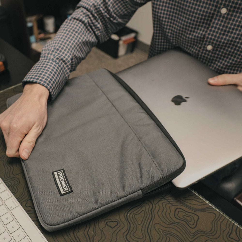 A person in a checkered shirt slides a laptop with an apple logo into the sleek gray Premier Body Armor Bulletproof Laptop Case on the desk. The partial keyboard and mouse pad view indicates a stylish workspace where safety meets style, featuring Level IIIA protection for peace of mind.