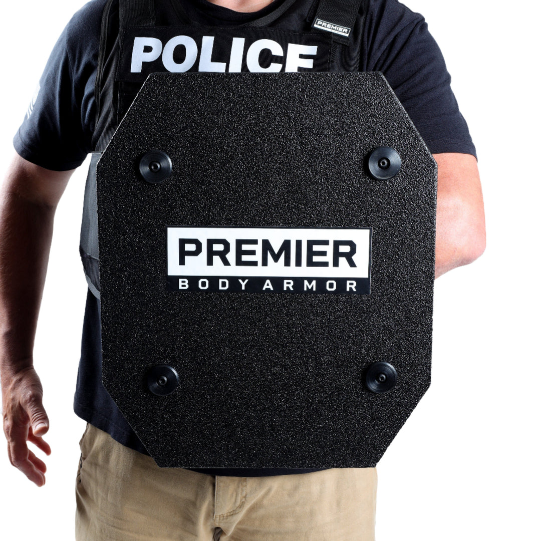 A person wearing a black vest marked "Police" and holding a lightweight defense shield labeled "STRATIS Pro Shield Level III+ by Premier Body Armor." The person is dressed in beige pants and a navy blue shirt. The background is white.