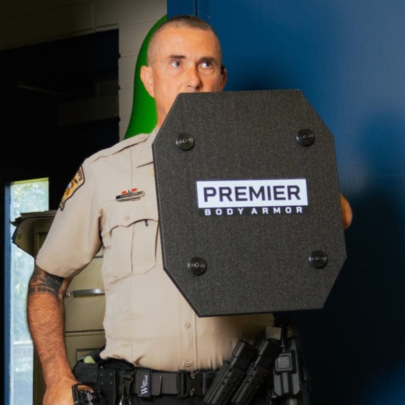 In a room with white tiled walls, a whiteboard, and a sink, a uniformed security officer stands in the doorway holding a STRATIS Pro Shield Level III+ by Premier Body Armor. The door behind him is open to darkness as he stays vigilant with rifle-rated protection.