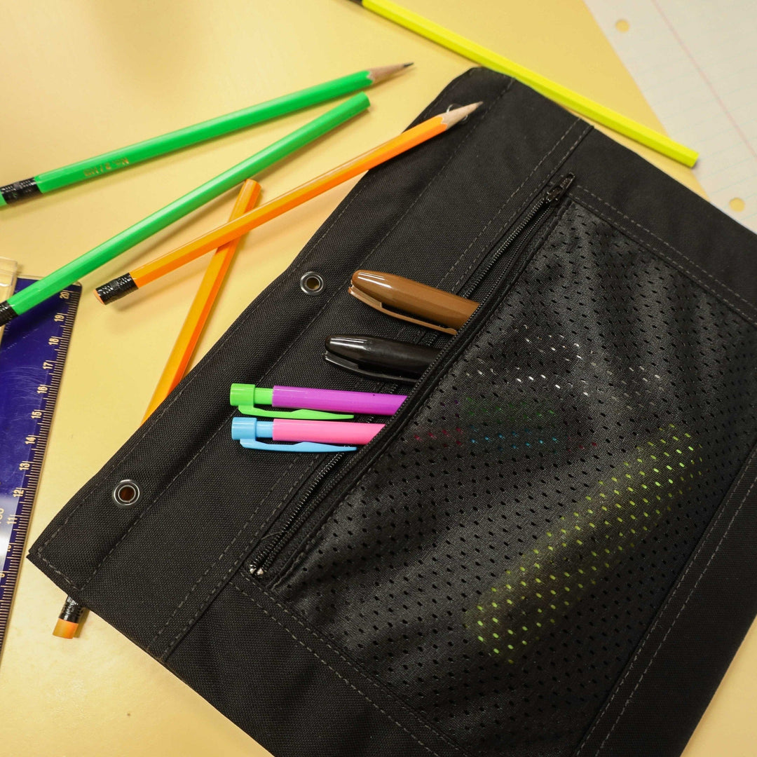 A Premier Body Armor Level IIIA 3-Ring Pencil Pouch, resembling sleek body armor, sits on a table with two pens inside. Nearby are three colored highlighters, a ruler, several pencils, and a piece of lined paper.