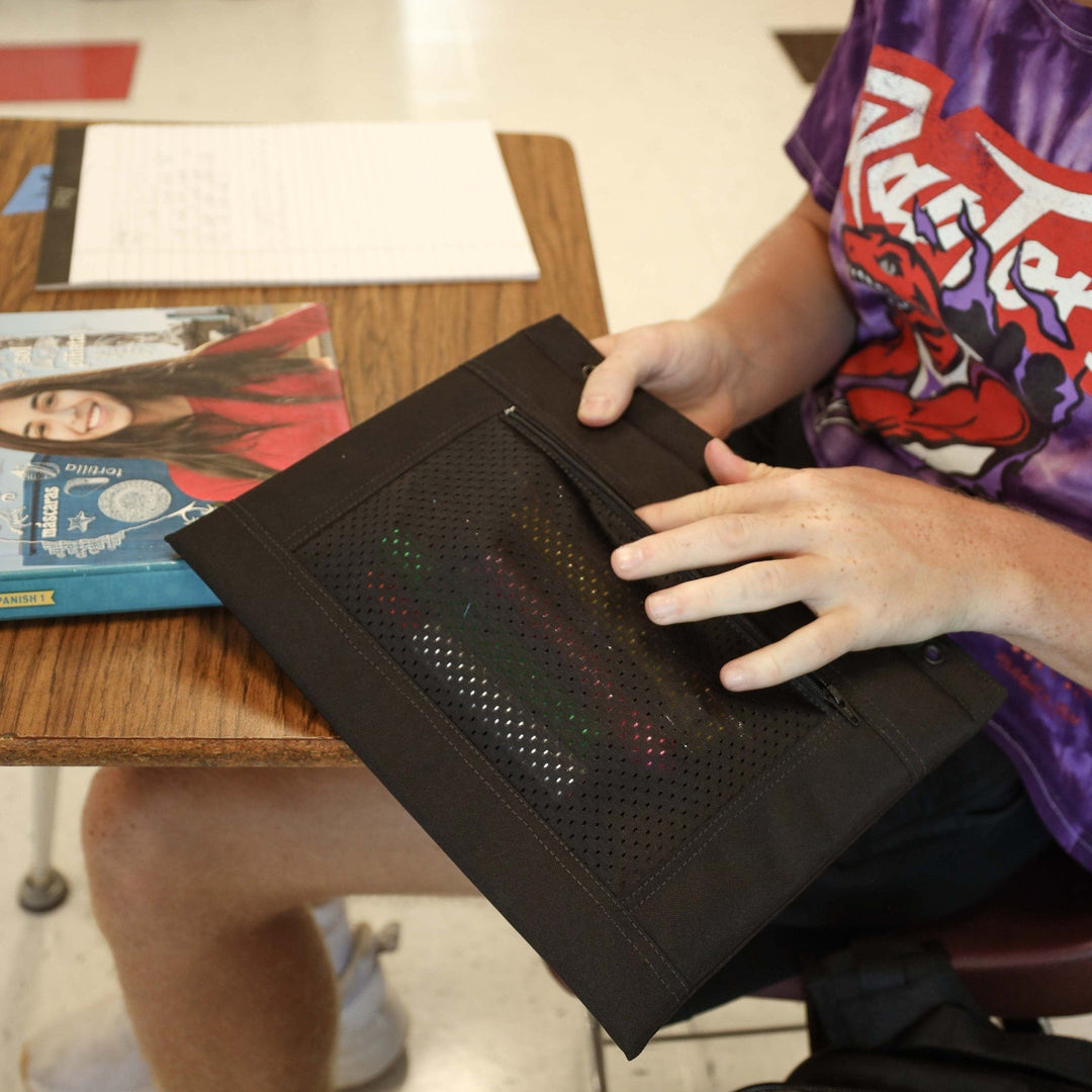 A person in a purple graphic t-shirt sits at a desk, touching the top of a Premier Body Armor Level IIIA 3-Ring Pencil Pouch. A magazine and a paper sheet are also on the desk, suggesting it’s a classroom setting.