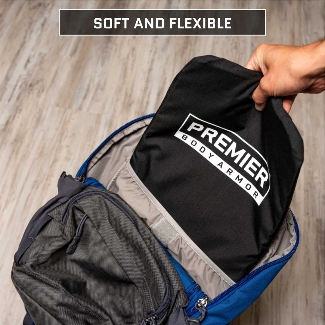 A hand is placing a "Premier Body Armor" 10.5x14 Level IIIA Insert with a CORDURA outer shell into a blue and gray backpack on a wooden floor. The insert, black with white text, slips easily into the open main compartment. A caption above reads "Soft and Flexible.