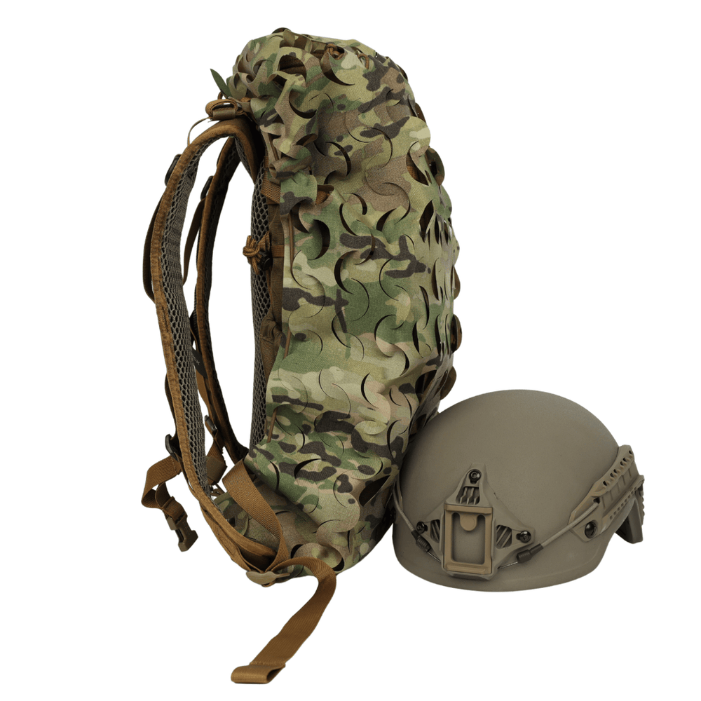 The OPE Gear Backpack Scrim - Camouflage Pack Cover by OPE Gear is shown standing upright beside a matte tan military helmet with mounting hardware, both set against a plain white background.