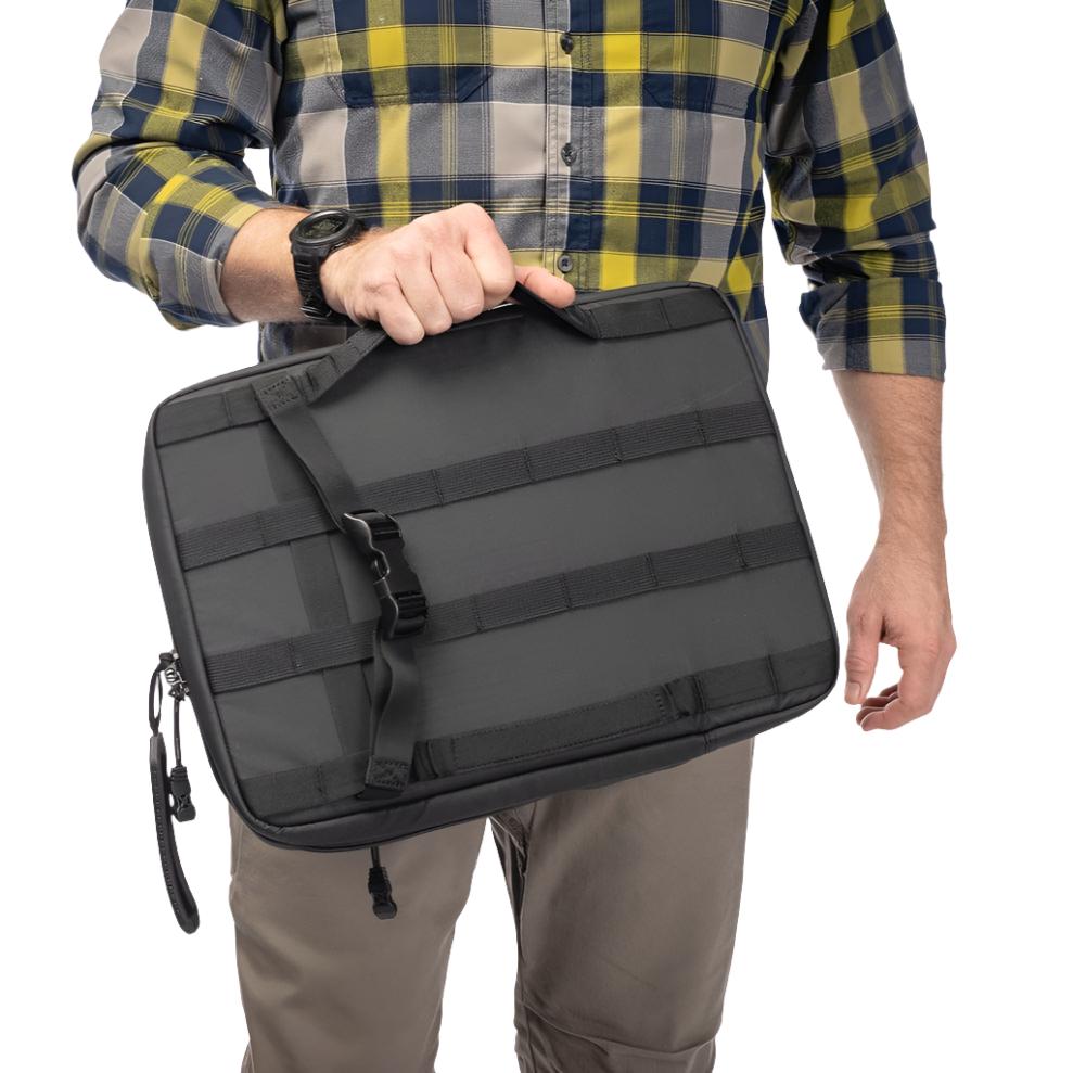 A person wearing a yellow and blue plaid shirt and khaki pants holds a Vertx VTAC Pistol Scabbard by its handle. The black tactical pouch, designed for firearm storage by Vertx, features molle webbing and a zipper closure. The person's face is not visible.