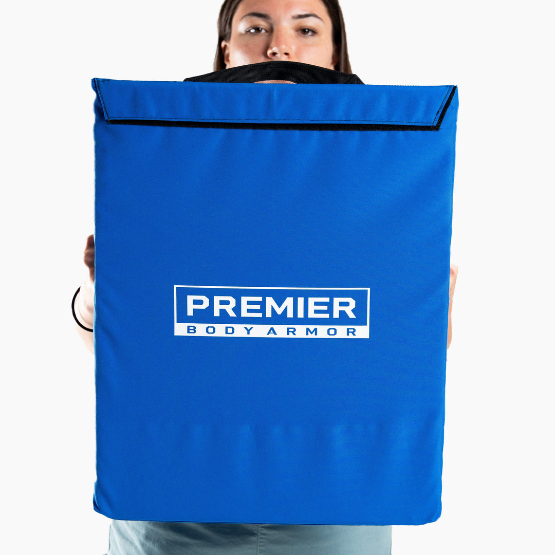 A person holds a blue panel with the text "Premier Body Armor" printed in white on it. The panel, part of a First Aid Shield, features a black handle at the top. The background is white, emphasizing its role in school safety and emergency response kits.