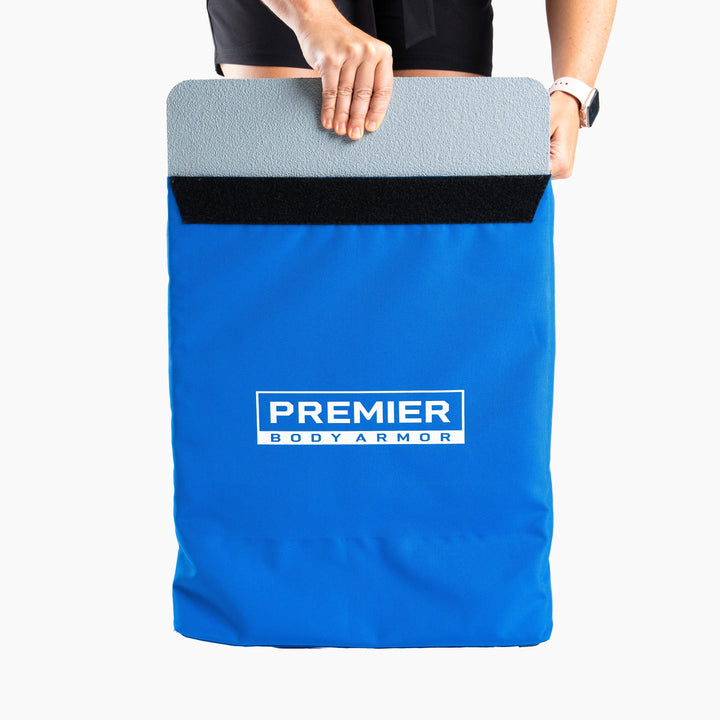 A person places a gray Level III+ First Aid Shield into a blue Premier Body Armor carrier with a Velcro strap. The person's hands are visible, wearing a black watch and black shorts. The brand name "Premier Body Armor" is prominently displayed on the carrier, ensuring school safety.