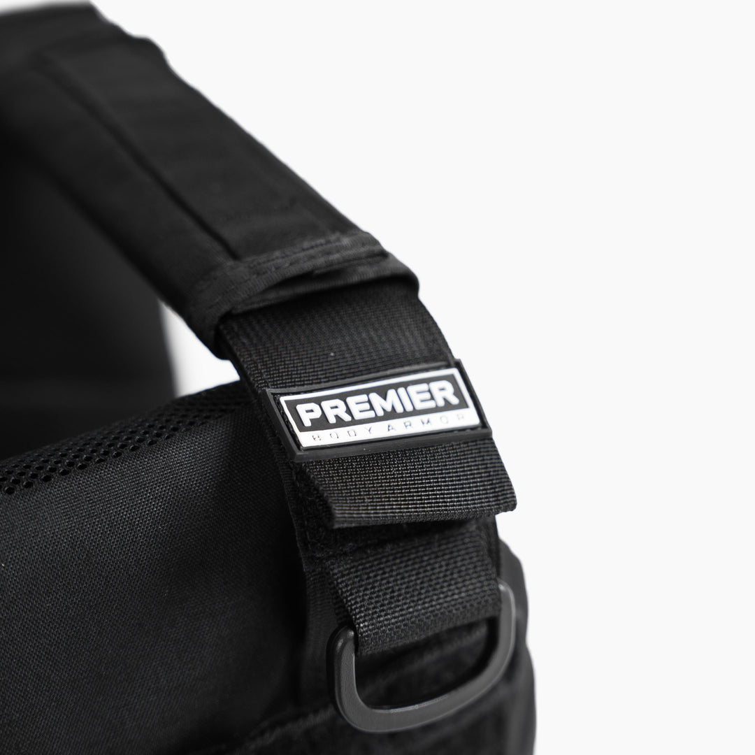 Close-up of a black strap with a "Premier Body Armor" metal label on the Falcon Carrier, a lightweight, low-profile plate carrier for 8x10 plates. A metal D-ring sits below the label against a plain white background.