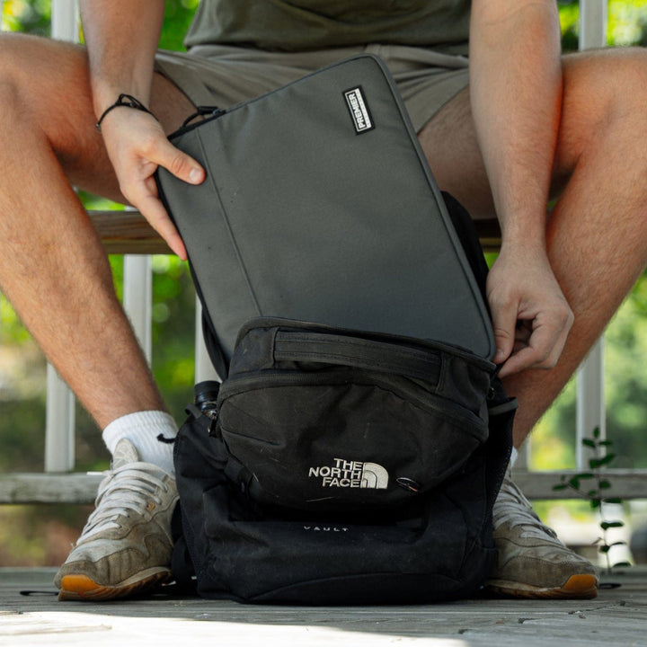 A person sits on a bench, placing a laptop into a Premier Body Armor Bulletproof Laptop Case before tucking it into a black North Face backpack. Wearing shorts, sneakers, and a wristband, they enjoy lush green scenery—a true EDC enthusiast.