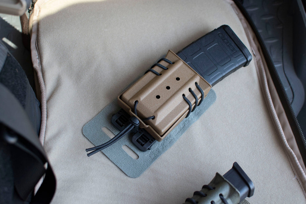 A single black magazine is secured in a brown polymer magazine pouch, which is lying on a beige fabric surface. The pouch is attached to the Vertx Molle Adaptor Panel - Double's gray mounting panel using black cords.