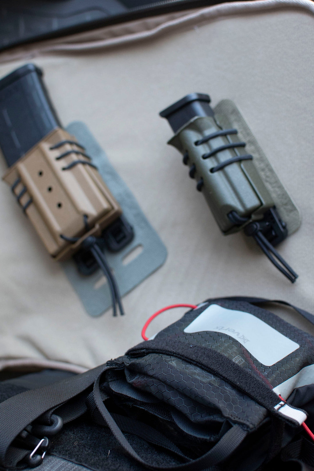 Close-up of the Vertx Molle Adaptor Panel - Single featuring two MOLLE-compatible rifle magazine pouches mounted on a fabric panel, one in brown and one in green. Below them is a black bag with hexagonal stitching, a visible patch, and red cord attached.