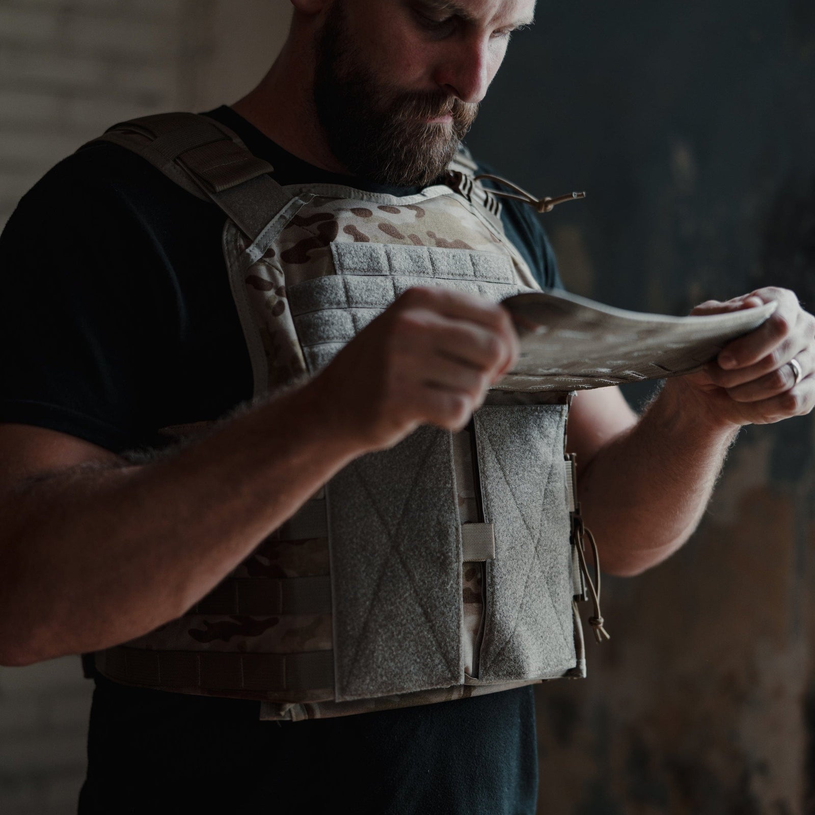 In a dimly lit room with textured walls, a bearded man examines a piece of fabric on his Core Mission Plate Carrier w/ Level IIIA Cummerbund Armor by Premier Body Armor. The tactical vest, equipped with MOLLE webbing, appears tailored for his core mission needs.