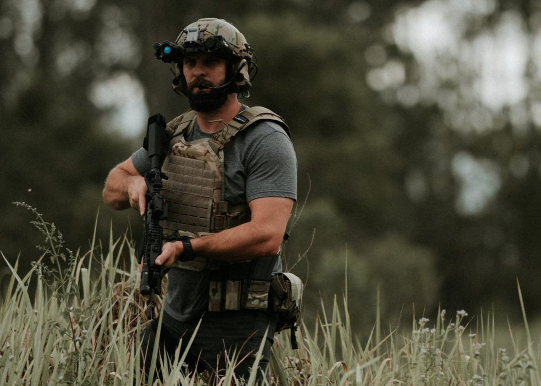 Man in tactical gear standing in a grassy field with trees in the background