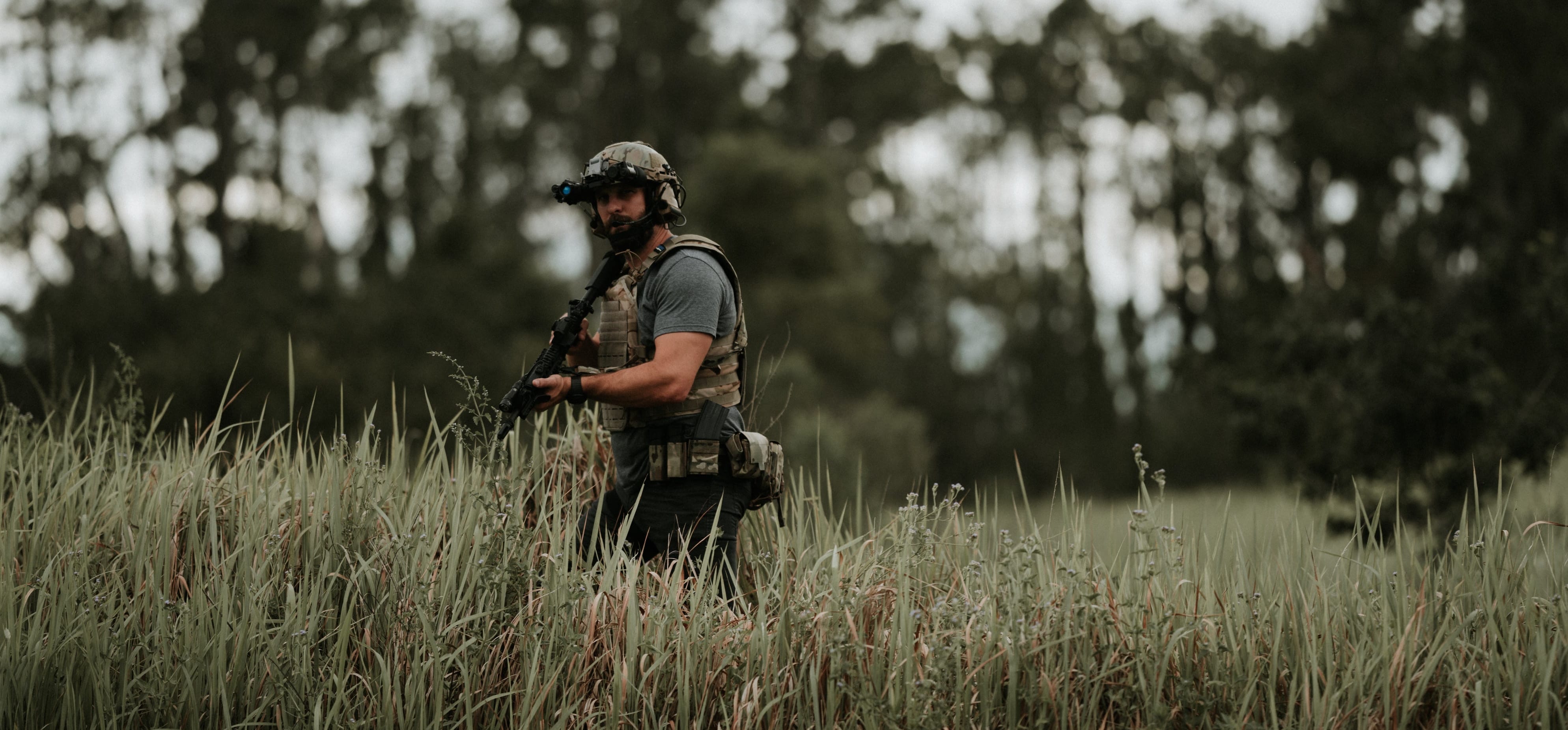 A person in tactical gear, including a helmet and body armor, stands in a tall grassy field holding a rifle, with a blurred background of trees and an overcast sky.