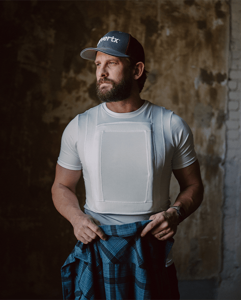 A man with a beard is wearing a baseball cap, a fitted white t-shirt with a padded vest over it, and holding a blue plaid shirt. He stands against a worn, textured background with his head turned slightly to the side.