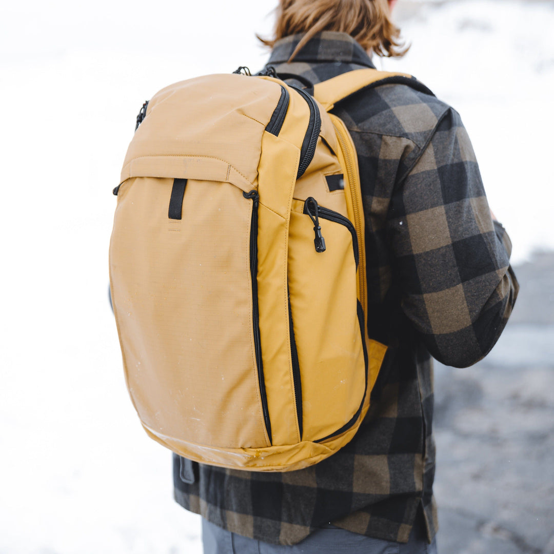 A person with long hair is wearing a checkered jacket and carrying a large yellow Armored Vertx Gamut 3.0 Bag Bundle by Vertx/Premier. The background appears to be a snowy outdoor area. This bag features multiple compartments and zippers for efficient storage.