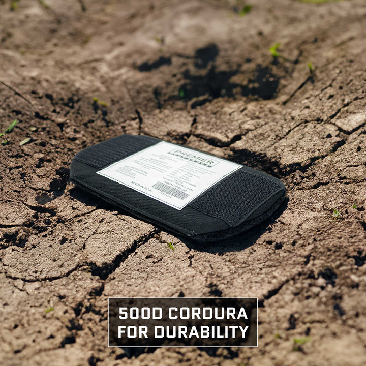 A black rectangular insert, branded as the Eberlestock Bando Bag Level IIIA Insert by Premier Body Armor, rests on cracked, dry soil. Text beneath reads "500D Cordura for Durability," emphasizing its strength against NIJ ballistic standards. Sparse green shoots appear in the background.