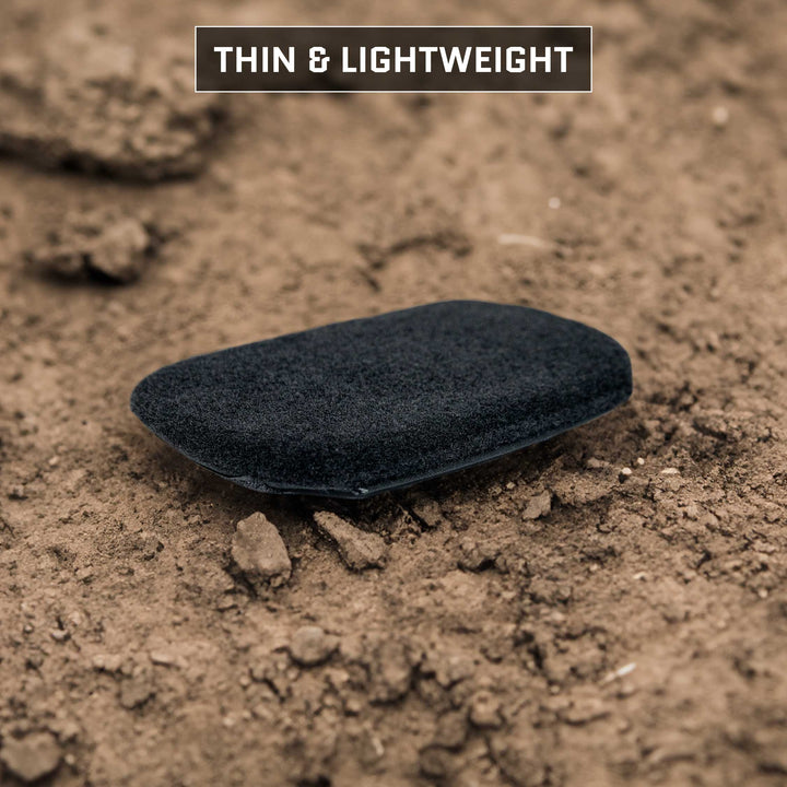 A thin, lightweight black object rests on dry, brown soil, reminiscent of the Eberlestock Bando Bag Level IIIA Insert by Premier Body Armor. The background features scattered rocks and uneven terrain. At the top, text reads “THIN & LIGHTWEIGHT.”