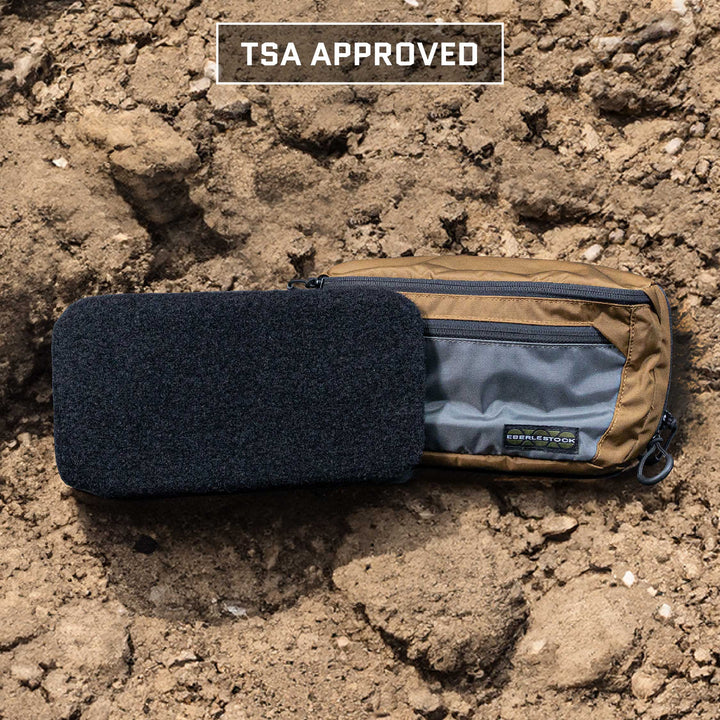 A khaki Eberlestock Bando Bag XL from Premier Body Armor, equipped with a Level IIIA insert for enhanced security, lies on the rocky ground alongside a black rectangular pouch. The bag is adorned with multiple zippers and displays a patch with text, showcasing its TSA-approved status.