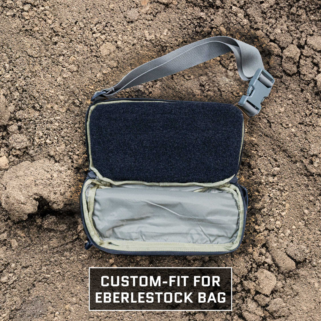 A gray and black custom-fit pouch with an open flap revealing an empty compartment is displayed on a dirt background. Text at the bottom reads "Custom-fit for Premier Body Armor Eberlestock Bando Bag XL Level IIIA Insert." A gray strap with a buckle is attached to the pouch, designed to easily accommodate Level IIIA Body Armor for added ballistic protection.