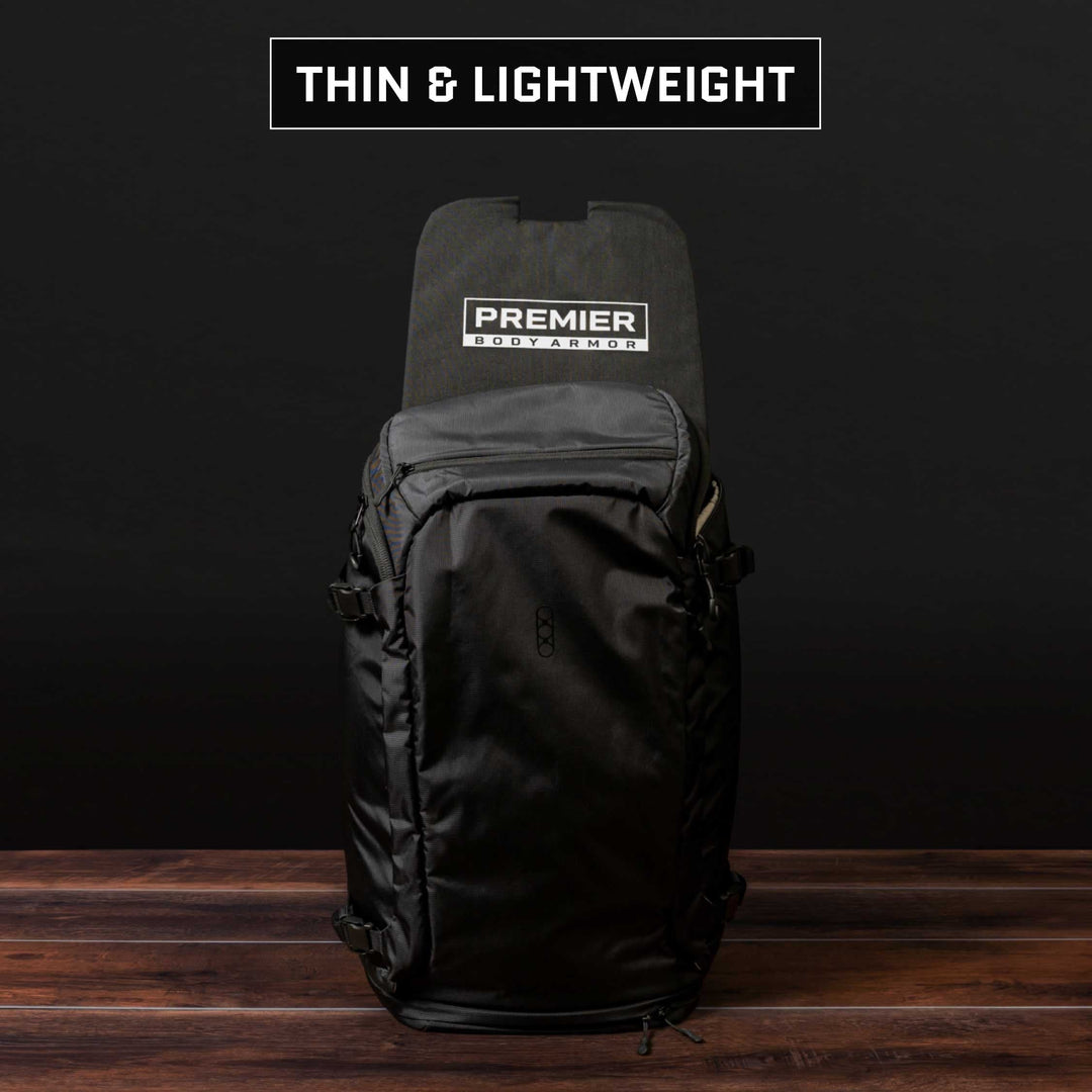 A black Eberlestock Fade Adapt Backpack with a Premier Body Armor Level IIIA Insert panel stands upright on a wooden surface. The words "Thin & Lightweight" appear at the top against a dark background, suggesting its TSA-approved convenience and sleek design.