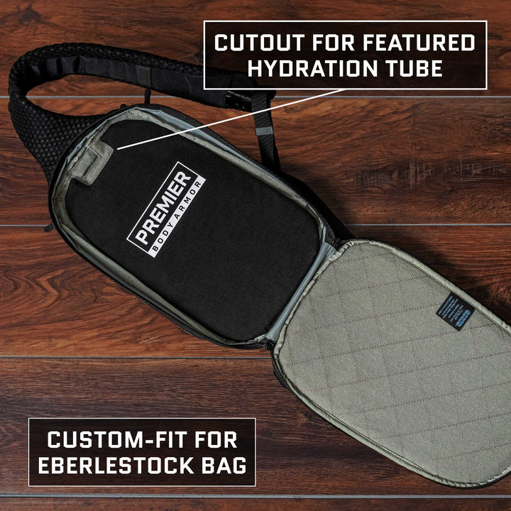 On a wooden surface, an open backpack reveals black protective padding featuring the "Premier Body Armor" branding. The product is described as having a cutout for a hydration tube and is designed to custom-fit the Eberlestock Fade Sling Bag, featuring TSA Approved Level IIIA Body Armor.