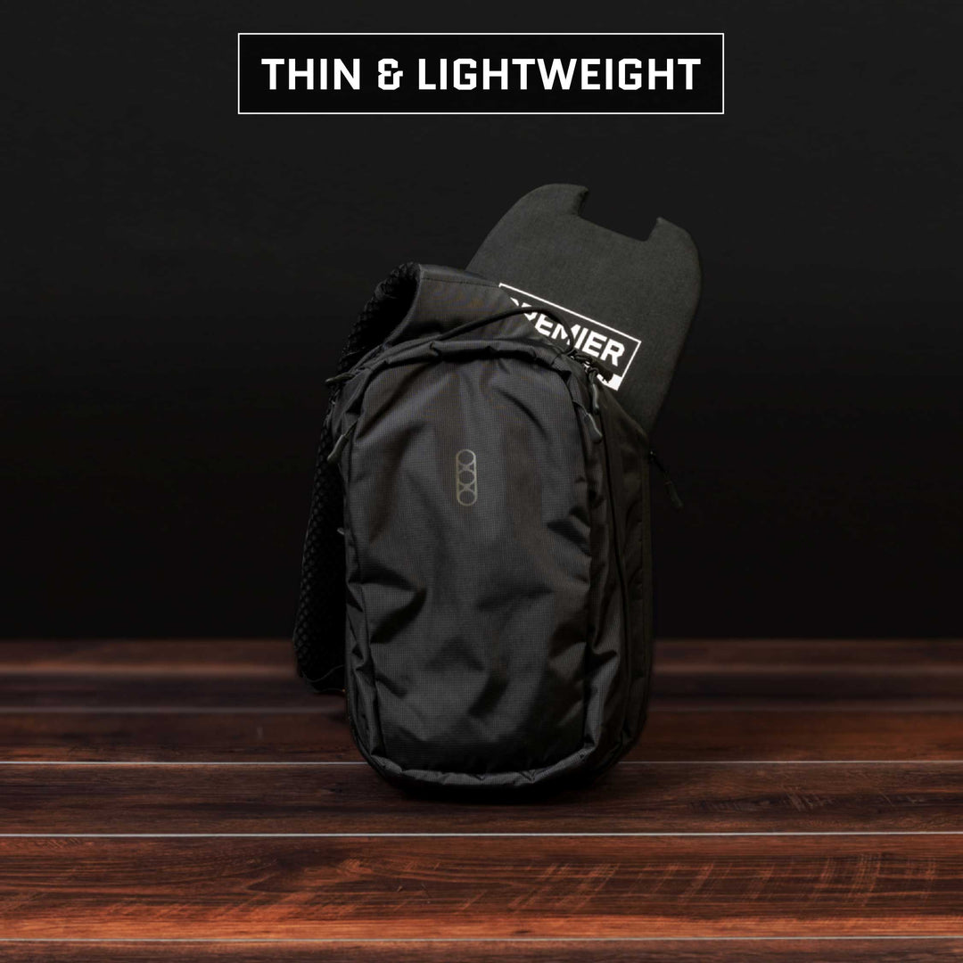 A black, thin, and lightweight Premier Body Armor Eberlestock Fade Sling Level IIIA Insert is set against a dark background. The bag is partially open, revealing a skateboard inside. Text at the top reads "THIN & LIGHTWEIGHT." The wooden surface beneath adds contrast to the scene.