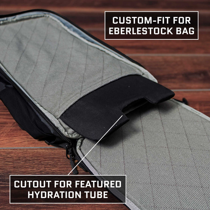 The image captures a close-up of an Eberlestock Fade Transport 21 Level IIIA Insert by Premier Body Armor, featuring quilted fabric and a partially open zipper. The text emphasizes its custom-fit design for Level IIIA Body Armor, complete with a hydration tube cutout. The scene is set against wooden flooring.