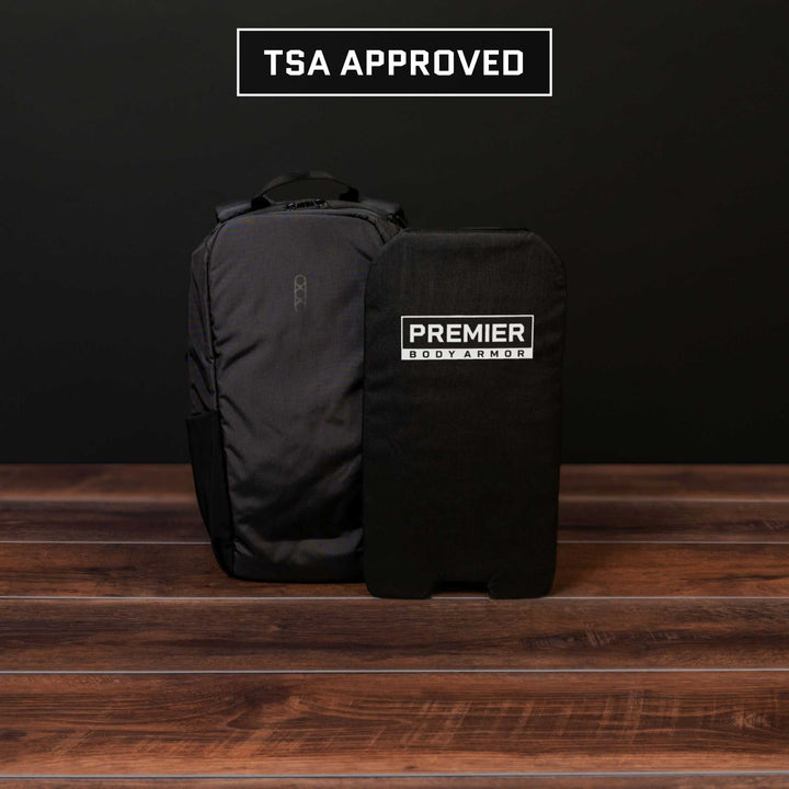 A black backpack showcasing the Eberlestock Fade Transport 21 with a Level IIIA Insert is displayed on a wooden surface against a dark background. The armor panel, bearing the "Premier Body Armor" label, is TSA approved and ideal for modern travel requirements.