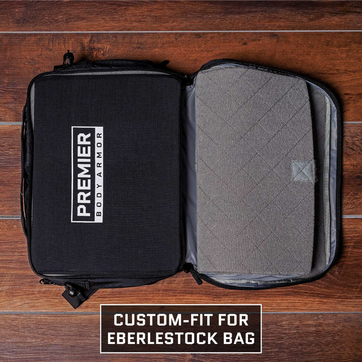 A Premier Body Armor case, in black, lies open within an Eberlestock Fade Briefcase on a wooden floor. The inside showcases a gray cushioned lining. At the bottom, the text states "Custom-fit for Eberlestock Bag, TSA Approved, and Level IIIA Body Armor.