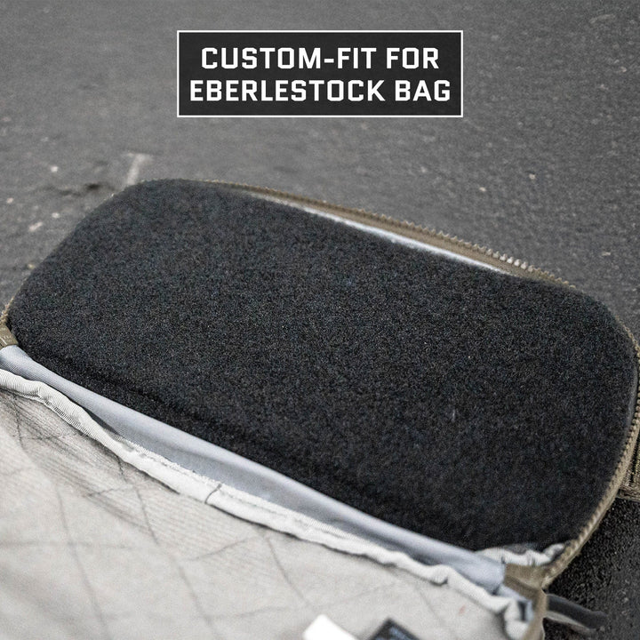 Close-up of the Eberlestock Fade Flex Level IIIA Insert by Premier Body Armor inside an Eberlestock bag, showcasing its custom-fit design. The open compartment reveals a dark fabric insert tailored to the bag's shape. Background is a textured surface with "Custom-Fit for Eberlestock Bag" in white text above.