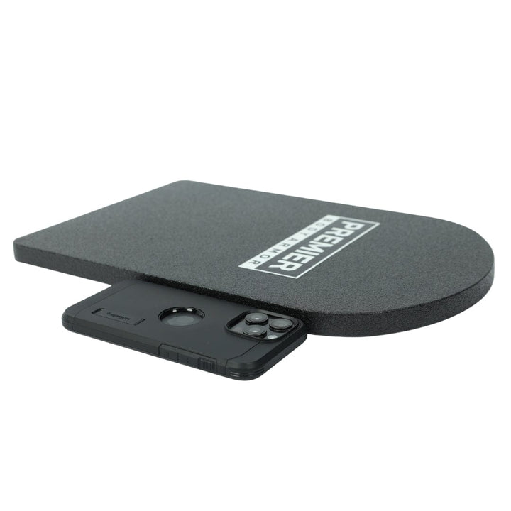 A thick, rectangular black 10x15 Special Threat Rifle-Rated Backpack Insert by Premier Body Armor with a rounded edge is displayed on a white background. A black smartphone in a bulky case sits in front for size comparison.