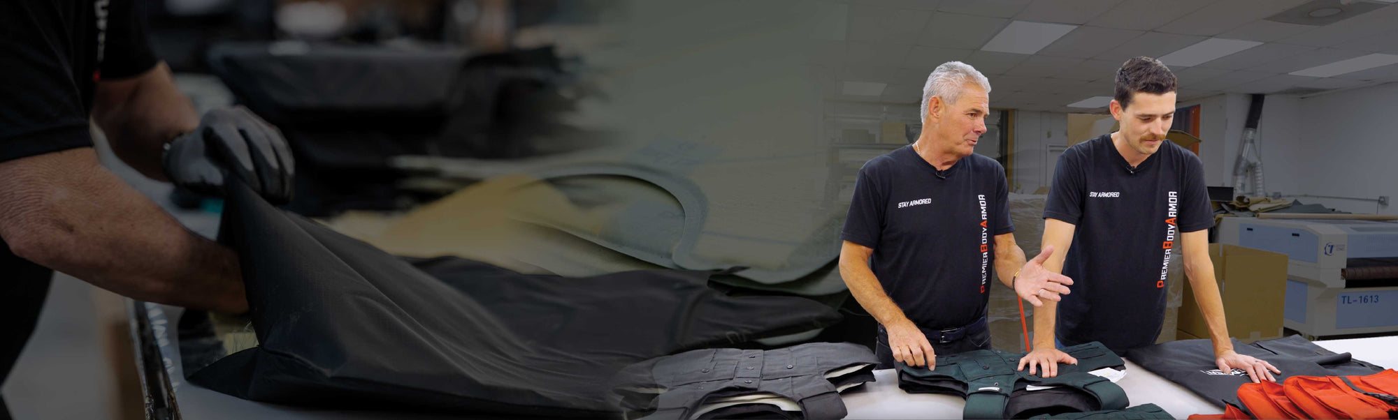 Two men wearing black shirts are examining fabric pieces laid out on a table in a brightly lit room. The man on the left gestures as he talks, while the man on the right looks at the materials. Another person works with fabric in the background.