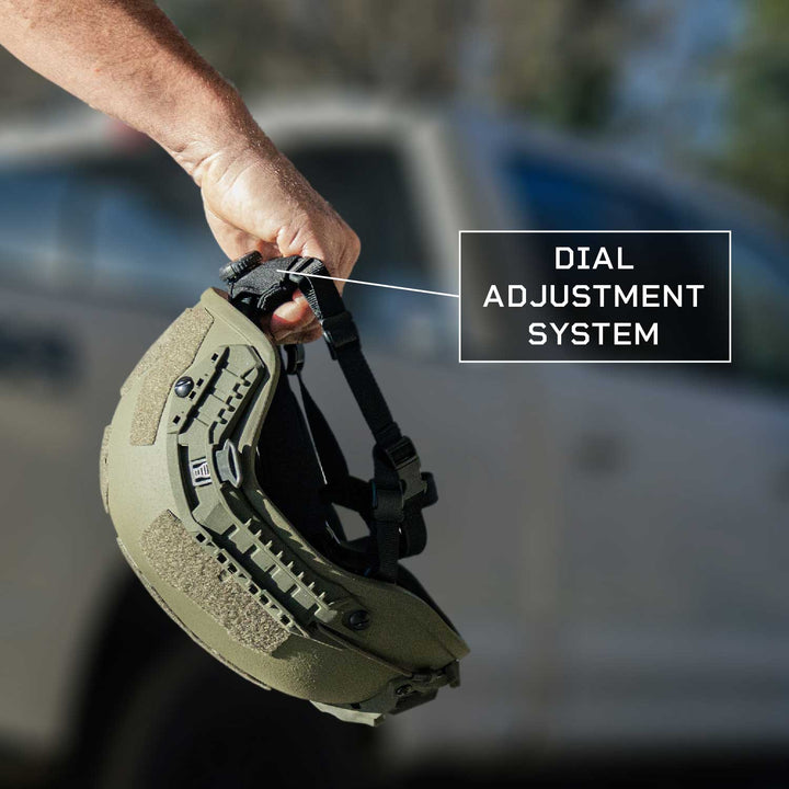 A hand holds a Fortis Ballistic Helmet by Premier Body Armor featuring a dial adjustment system and straps. The lightweight helmet is olive green, with a blurred vehicle in the background highlighting advanced protective technology.