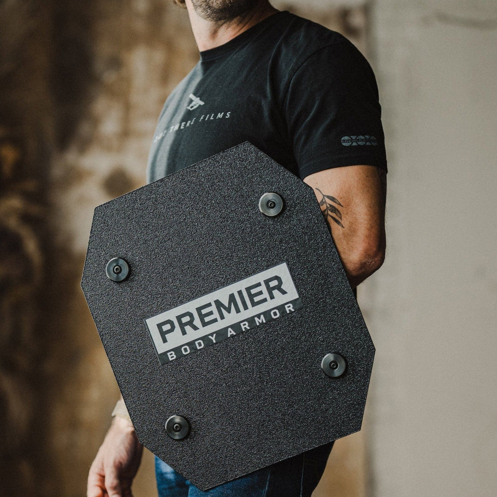 A man in a cap and black t-shirt holds a Premier Body Armor Home Shield, standing with a black dog on wooden floors against a textured wall, showcasing top home protection.